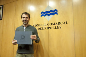 El president, Joaquim Colomer, mostrant un exemplar del llibre, davant la paret de la sala de plens on figura el logotip del consell comarcal