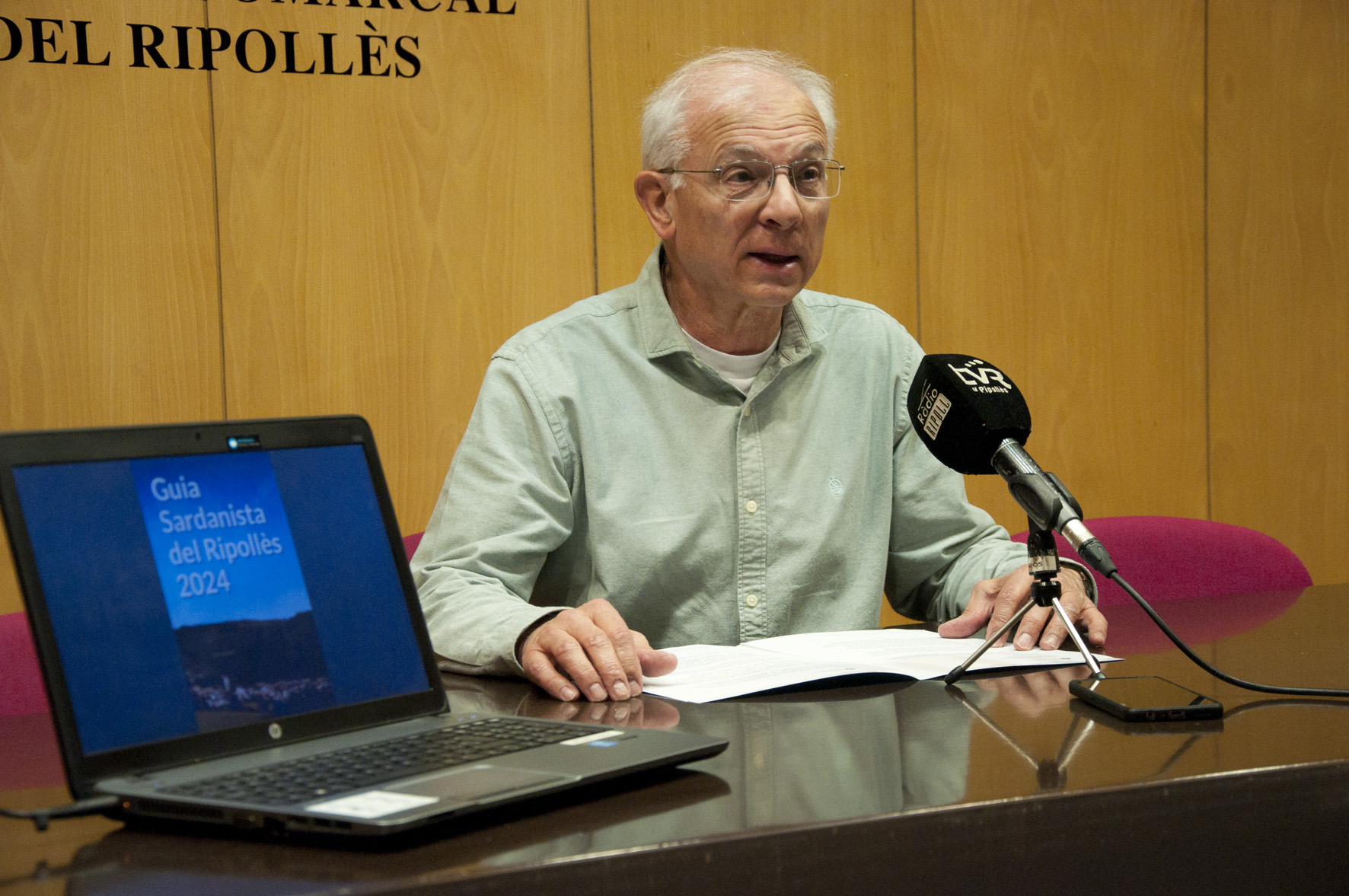 Amadeu Rosell, a la sala de plens, presentant la Guia sardanista del Ripollès 2024, que es mostra en un ordinador a la seva dreta.